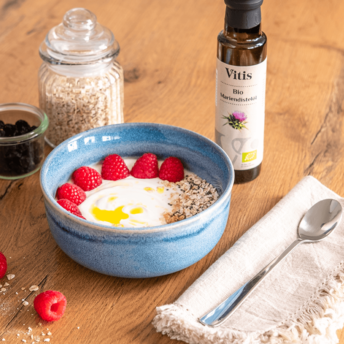 Eine Flasche Bio Mariendistelöl von Vitis steht neben einer blauen Schüssel Joghurt mit Müsli. Als Dekoration sind Himbeeren daneben gelegt.