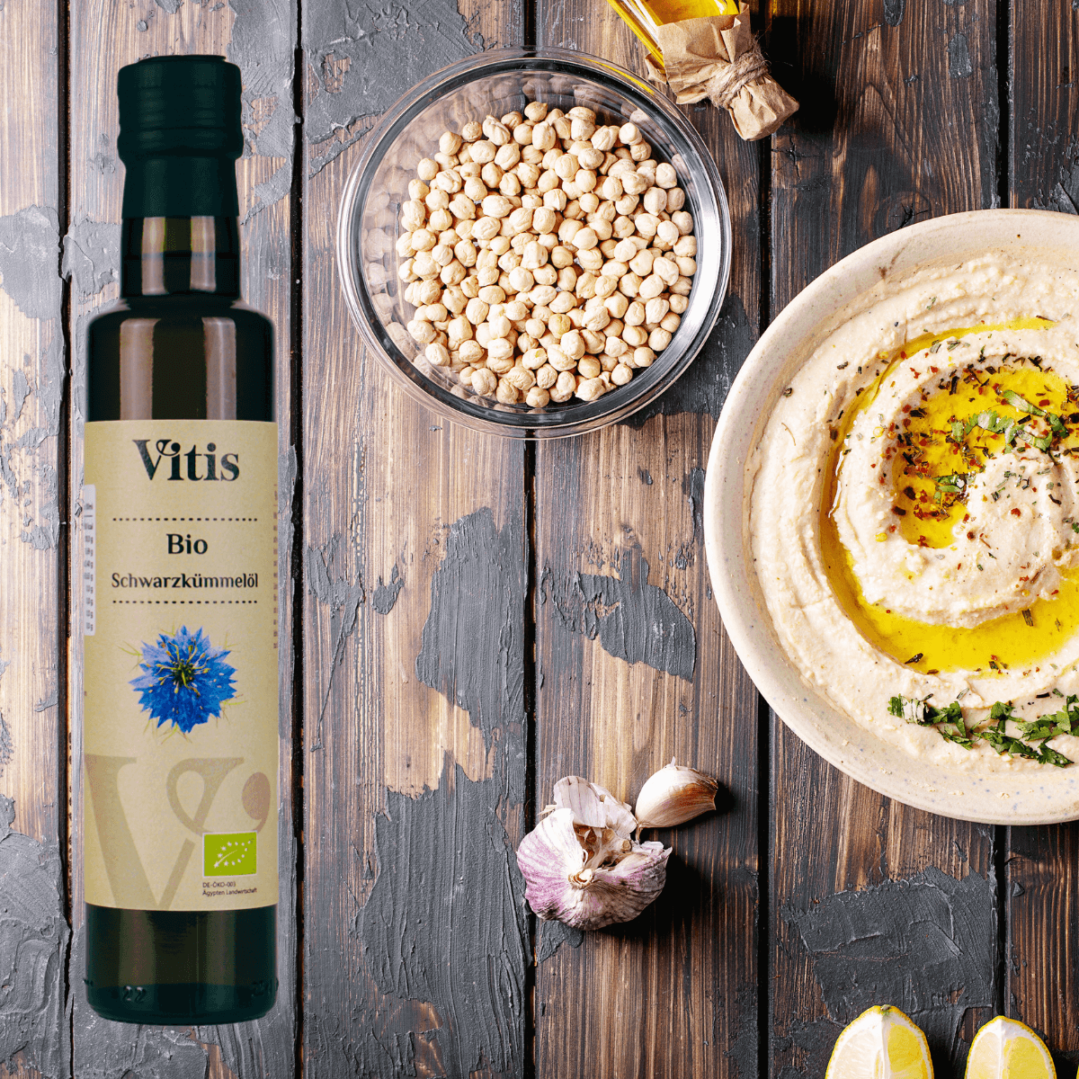 Eine Flasche Bio Schwarzkümmelöl von Vitis liegt neben einer Schüssel Humus. Als Dekoration ligen Knoblauch und Zitrone daneben.