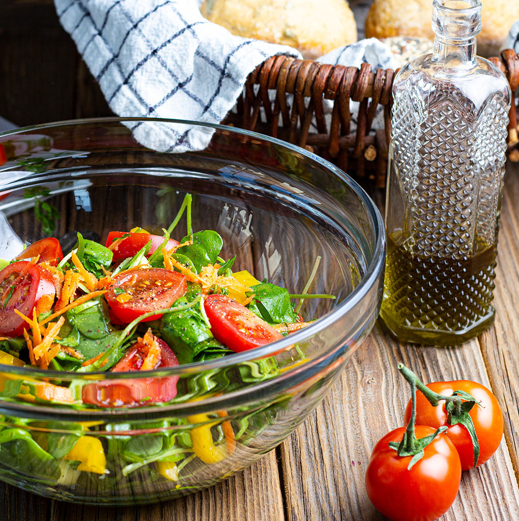 eine Schüssel mit buntem Salat steht vor einem Brotkorb neben einer hübschen Karaffe mit dunkelgrünem Öl und zwei Tomaten