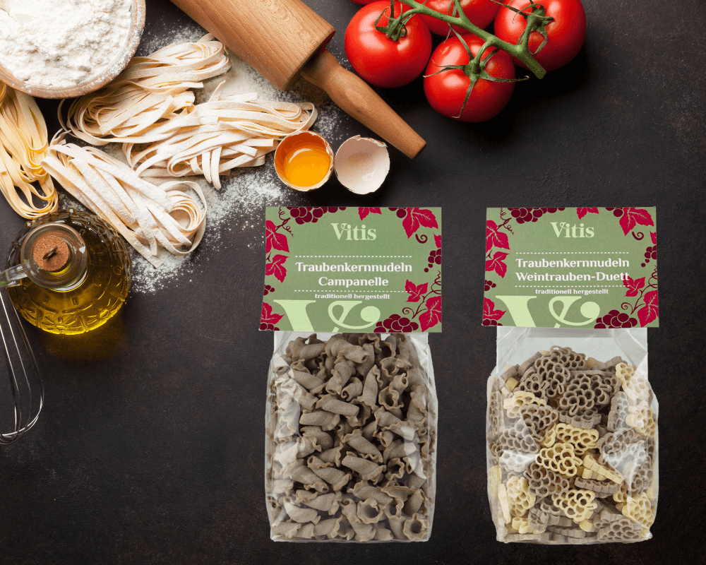 Die Traubenkernnudeln Campanelle und Weintrauben Duett auf dunklem Hintergrund. Auf der linken Seite ist Pasta in verschiedenen Formen und Farben zu sehen.
