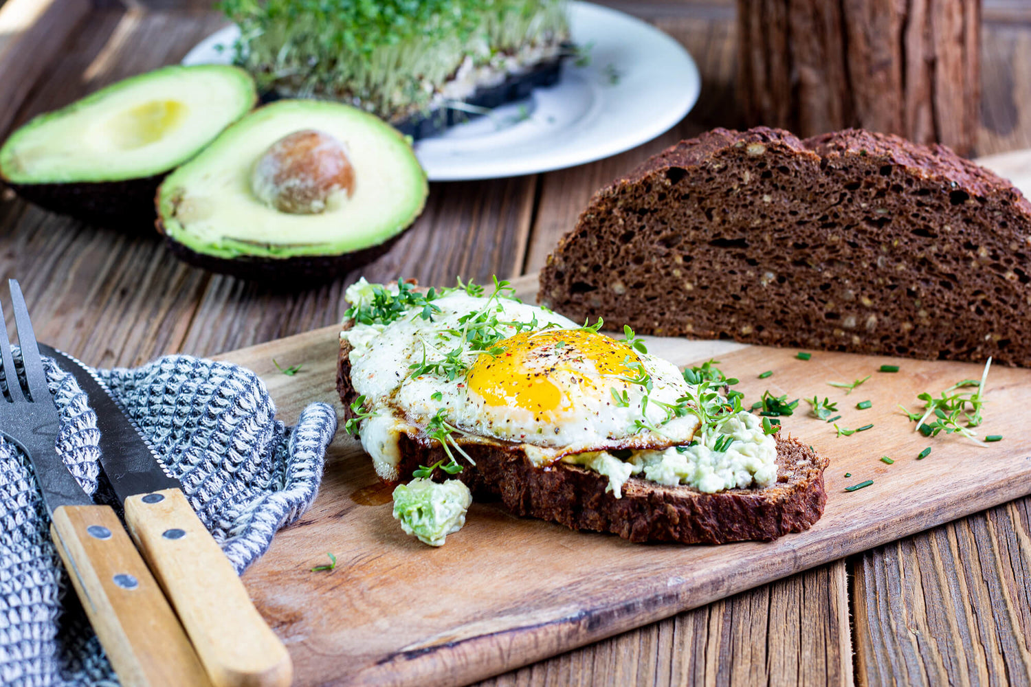 Ein Traubenkernbrot mit einer abgeschnittenen Scheibe im Vordergrund. Auf dem Traubenkernbrot ist ein Spiegelei. Als Dekoration ist Avocado-Hälften im Hintergrund.